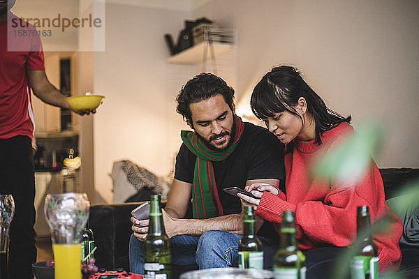 Männliche und weibliche Freunde benutzen ein Smartphone  während sie sich zu Hause ein Spiel ansehen