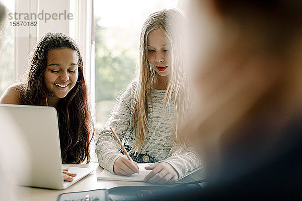 Studentin schreibt in Buch  während sie mit einem Freund am Tisch sitzt