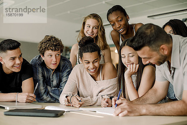 Lächelnder Lehrer erklärt Teenagern  während er sich im Klassenzimmer über den Tisch beugt