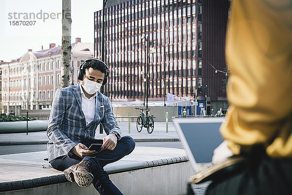 Geschäftsmann mit Gesichtsmaske sitzt draußen und benutzt ein Smartphone