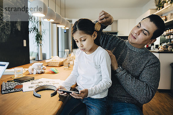 Vater kämmt der Tochter die Haare  während das Mädchen zu Hause am Tisch mit dem Handy telefoniert