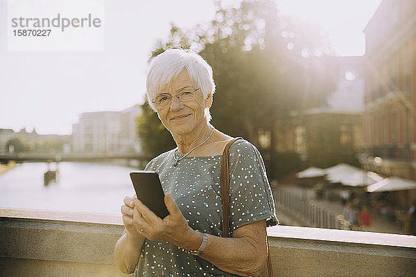 Porträt einer älteren Frau  die ein Mobiltelefon in der Hand hält und sich in der Stadt gegen ein Geländer stellt