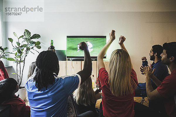 Fröhliche Freunde mit erhobenen Armen beim Fußballspiel im Wohnzimmer