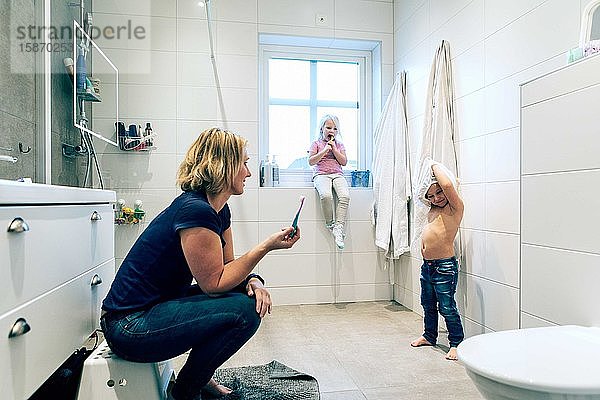 Seitenansicht der Mutter mit Blick auf die Tochter  die das Oberteil abnimmt  während das Mädchen am Fenster im Badezimmer sitzt