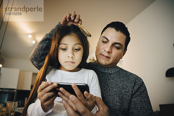 Niedriger Blickwinkel auf den Vater  der die Haare der Tochter kämmt  während das Mädchen zu Hause ein Mobiltelefon benutzt