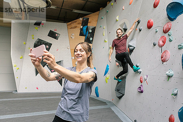 Lächelnde Frau  die sich mit einem männlichen Freund über ein Smartphone selbstständig macht  klettert im Fitnessstudio an einer Wand