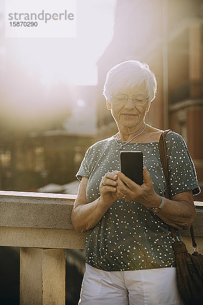 Ältere Frau benutzt Smartphone  während sie an einem sonnigen Tag in der Stadt am Geländer steht