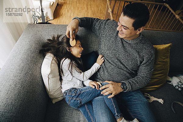 Hochwinkelansicht eines Vaters  der die Haare seiner Tochter kämmt  während er zu Hause auf dem Sofa sitzt