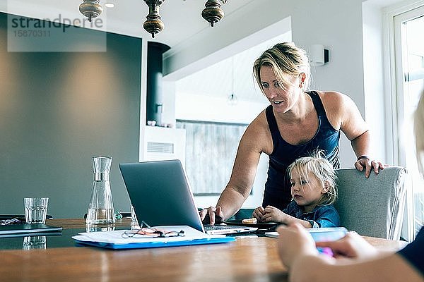 Mutter benutzt Laptop  während die Tochter zu Hause im Wohnzimmer sitzt