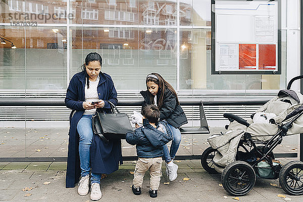 Mutter benutzt Smartphone  während sie mit Kindern an einer Bushaltestelle in der Stadt sitzt