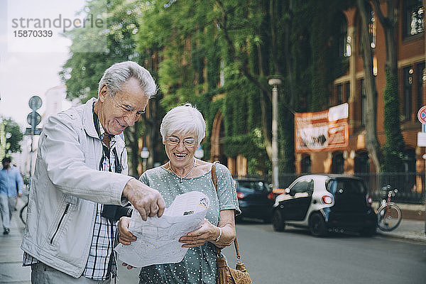 Lächelndes älteres Paar liest Karte  während es im Urlaub auf dem Bürgersteig in der Stadt steht