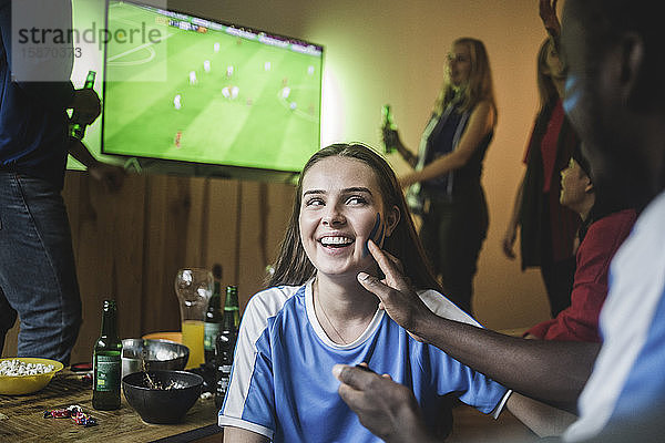 Mann trägt Gesichtsfarbe auf die Wange einer Frau auf  während er zu Hause ein Fußballspiel sieht