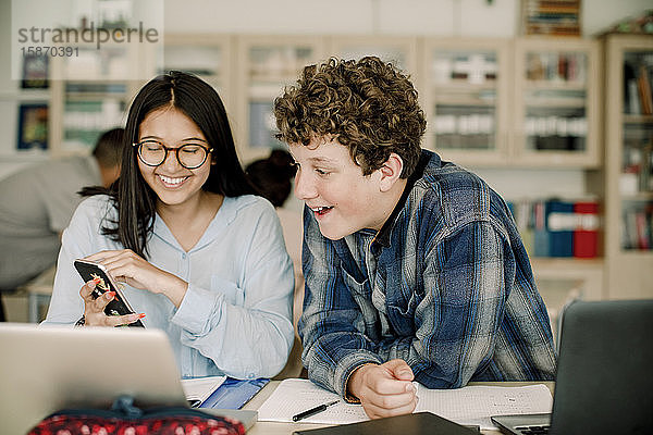 Lächelndes Teenager-Mädchen zeigt einem männlichen Freund ein Smartphone  während sie im Klassenzimmer sitzt