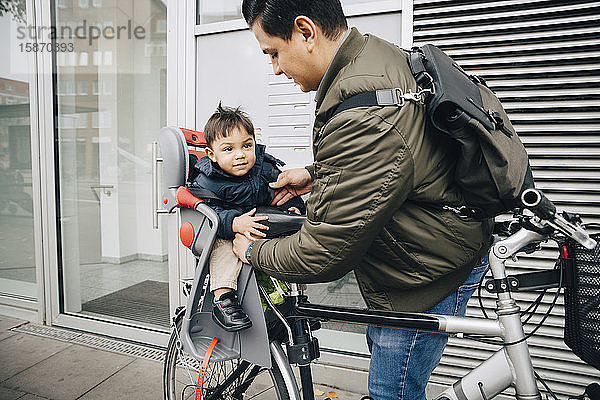 Vater hält Sohn im Sicherheitssitz eines Fahrrads auf dem Bürgersteig in der Stadt