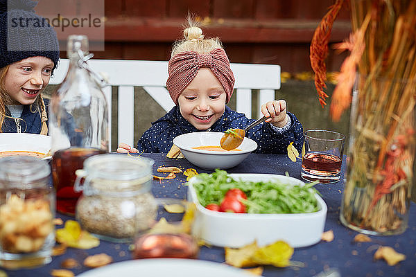 Lächelndes Mädchen isst vegetarische Suppe  während es neben einem weiblichen Geschwisterkind am Tisch sitzt