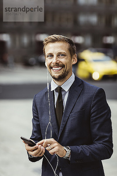 Porträt eines lächelnden Unternehmers  der ein Smartphone über In-Ear-Kopfhörer benutzt  während er im Freien steht
