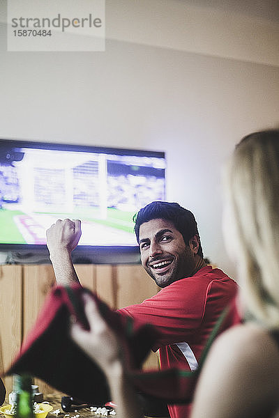 Lächelnder junger Mann schaut eine Freundin an  während er sich zu Hause ein Fußballspiel ansieht