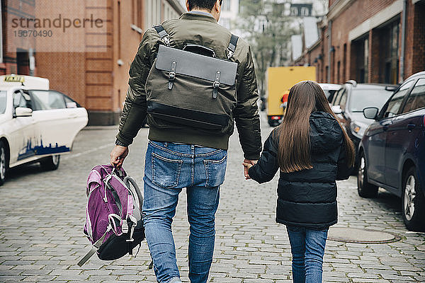 Rückansicht von Vater und Tochter mit Rucksack beim Spaziergang auf einem Fußweg in der Stadt