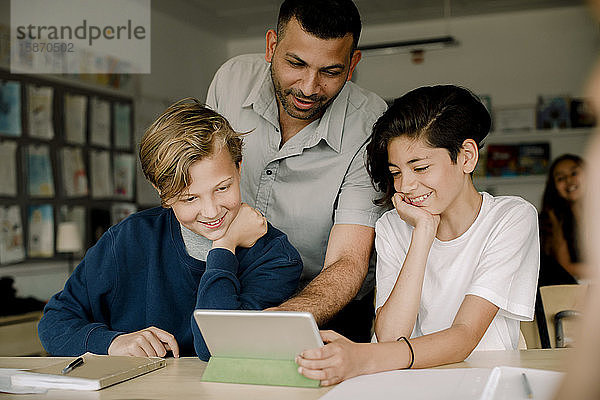 Lächelnder männlicher Tutor mit Schülern  die ein digitales Tablet benutzen  während sie im Klassenzimmer sitzen