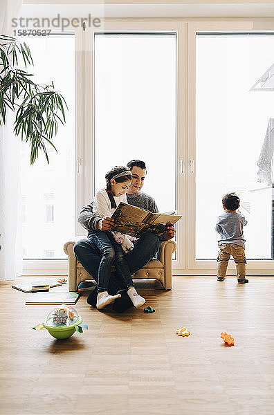 Der Vater liest der Tochter ein Buch in voller Länge vor  während der Sohn zu Hause am Fenster spielt