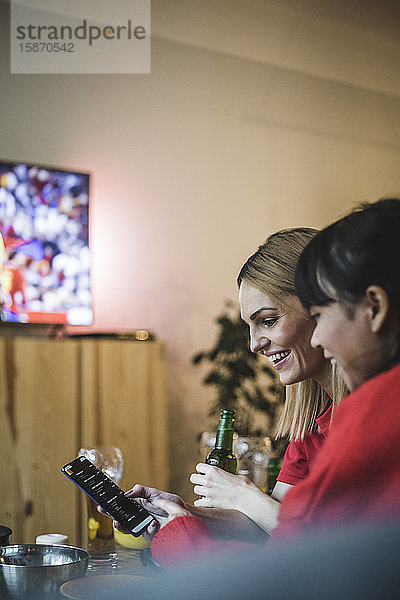 Lächelnde Freundinnen benutzen ein Smartphone  während sie zu Hause Fußball schauen