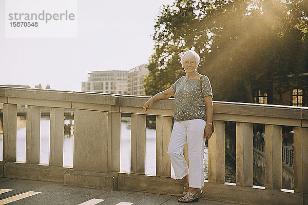 Porträt einer älteren Frau in voller Länge  die auf einer Brücke gegen ein Geländer in der Stadt steht