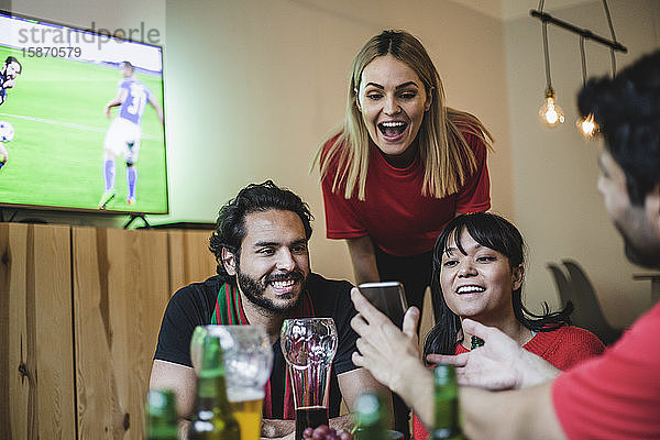 Lächelnde Freunde schauen auf ein Smartphone  während sie im Wohnzimmer Fußball schauen