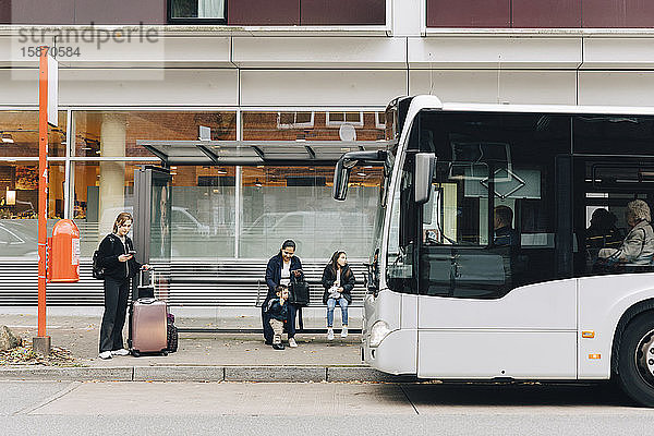 Frau in voller Länge mit Gepäck und Familie wartet an der Bushaltestelle in der Stadt auf den Bus