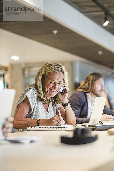 Lächelnde Geschäftsfrau schreibt Tagebuch  während sie im Büro am Smartphone telefoniert