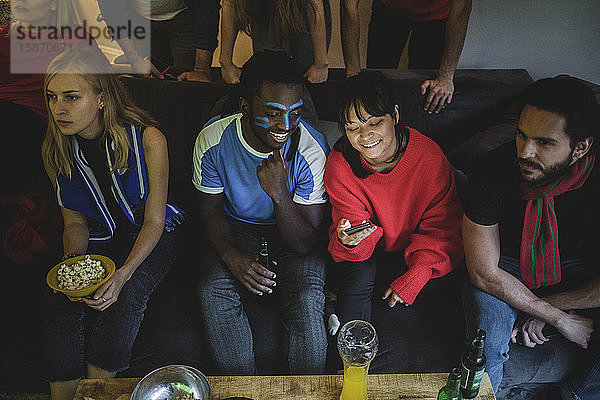 Männer und Frauen benutzen Smartphones  während sie mit Freunden zu Hause auf dem Sofa sitzen