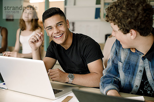 Lächelnde männliche Studenten  die den Laptop über dem Tisch benutzen  während sie im Klassenzimmer sitzen