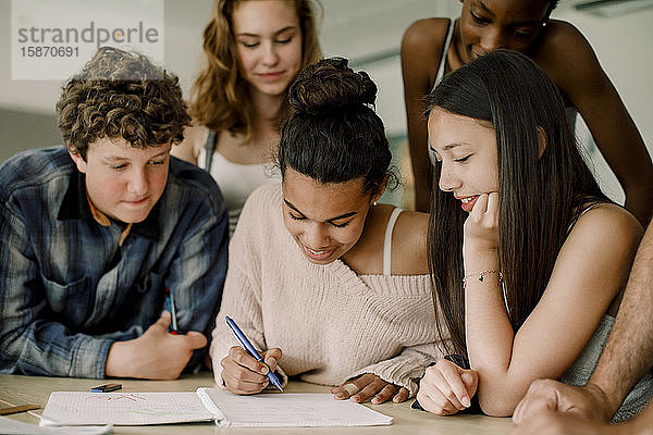 Lächelndes Teenagermädchen schreibt in Buch  während es inmitten von Freunden im Klassenzimmer sitzt