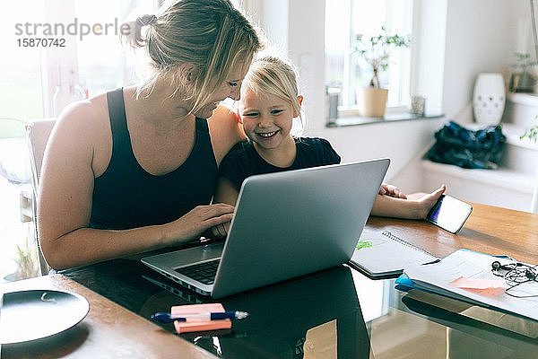 Tochter schaut auf den Laptop  während sie bei der Mutter zu Hause sitzt