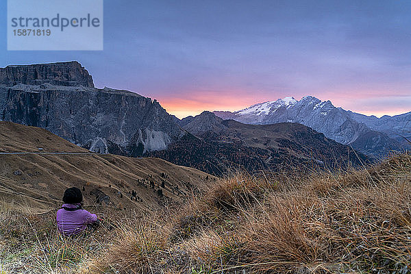 Rückansicht einer Frau  die im Gras sitzt und den Sonnenaufgang auf der Marmolada und dem Sass Pordoi  Sellajoch  Dolomiten  Südtirol  Italien  Europa  bewundert