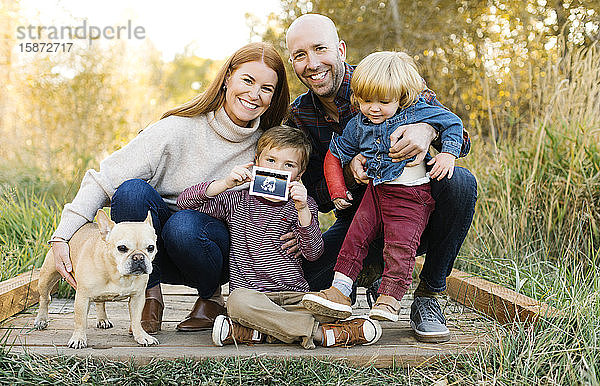 Lächelnde Familie mit Ultraschallfoto und Hund auf der Waldpromenade
