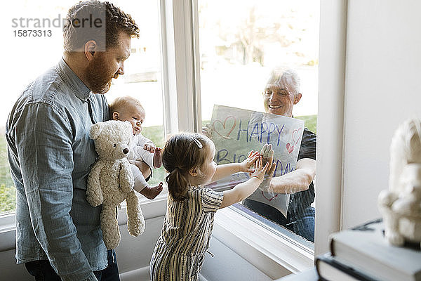 Großvater besucht Familie mit Enkelkindern (2-3 Monate  2-3) und zeigt Happy Birthday Zeichen durch das Fenster