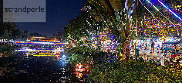 Eine traditionelle Holzbrücke über den Fluss Siem Reap  die zum Nachtmarkt des Siem Reap Art Centre führt  ist beleuchtet und mit Neonlichtern dekoriert. Auf dem Markt werden Souvenirs  Kunstwerke und Schnitzereien verkauft. Der Markt