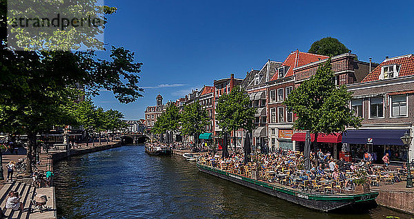 Stadt Leiden  Provinz Südholland  Niederlande  am Kanal 'Nieuwe Rijn'  die flachen Boote  die festgemacht sind  verwandeln sich in ein Café-Restaurant  in dem die Menschen die Sonne genießen. Die Stadt Leiden ist bekannt für ihre weltliche Architektur  ihre Kanäle  ihre Universität von 1590  die Geburtsstadt von Rembrand  die Stadt  in der im 16. Jahrhundert die erste Tulpe in Europa blühte.