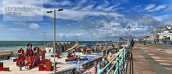 Großbritannien  East Sussex  Südküste Englands  Stadt Brighton und Hove  Foto von der Strandpromenade und dem Strand von Brighton an einem sonnigen Tag  mit Touristen  die an der Strandpromenade und auf dem Spielplatz spazieren gehen  Brighton liegt an der Südküste Englands und gehört zur Stadt Brighton und Hove. Die Lage von Brighton hat die Stadt zu einem beliebten Reiseziel für Touristen gemacht und ist das beliebteste Badeziel im Vereinigten Königreich für Touristen aus Übersee  das auch als die angesagteste Stadt des Vereinigten Königreichs bezeichnet wird.