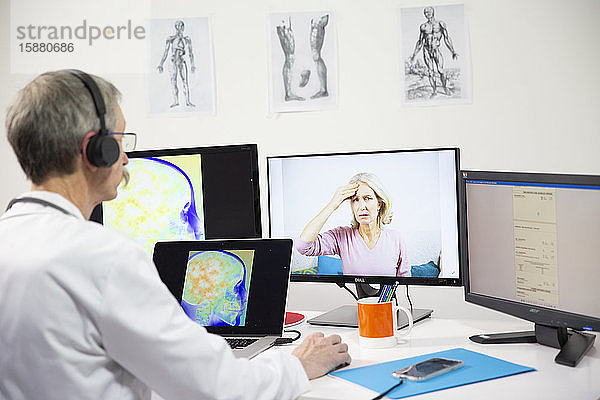 Ein Allgemeinmediziner während einer Videokonsultation mit einer 45-jährigen Frau wegen Kopfschmerzen. Auf dem Bildschirm ist eine Röntgenaufnahme ihres Gehirns zu sehen.