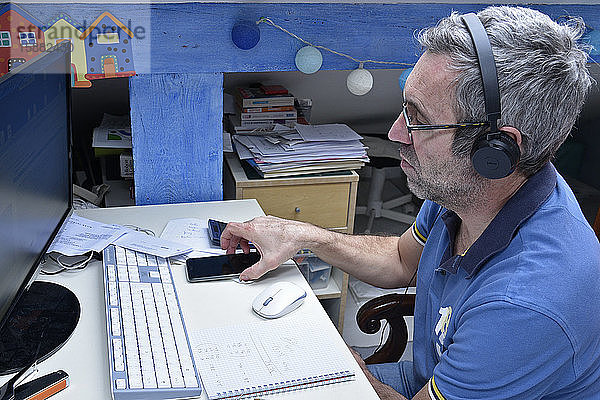 Mann bei Telearbeit zu Hause an seinem Schreibtisch mit Computer  Smartphone und Kopfhörern.