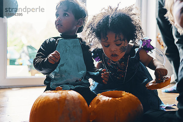 Zwei für Halloween verkleidete kleine Kinder  die mit zwei Kürbissen auf dem Boden knien.