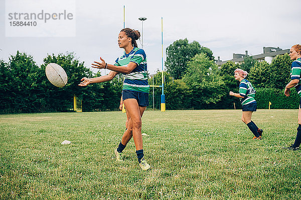 Drei Frauen in blauen  weißen und grünen Rugbyhemden auf einem Trainingsplatz  eine Frau gibt einen Rugbyball ab.