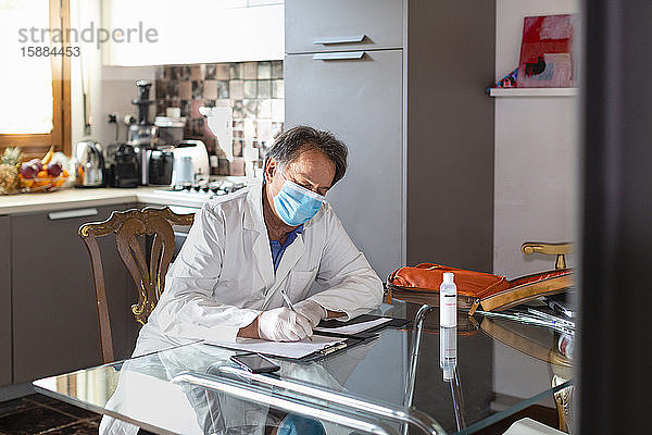 Arzt mit weißem Kittel  Gesichtsmaske und Schutzhandschuhen sitzt an einem Küchentisch und schreibt medizinische Notizen.