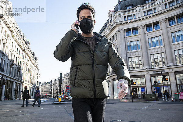 Ein Mann  der eine Gesichtsmaske und Plastikhandschuhe trägt  geht die Oxford Street entlang und telefoniert mit einem Mobiltelefon.