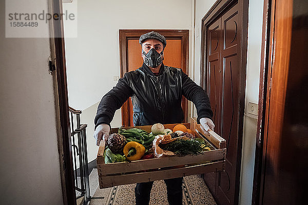 Mann mit flacher Kappe und Gesichtsmaske  der während der Corona-Virus-Krise in einem Wohnhaus eine Gemüsekiste lieferte.