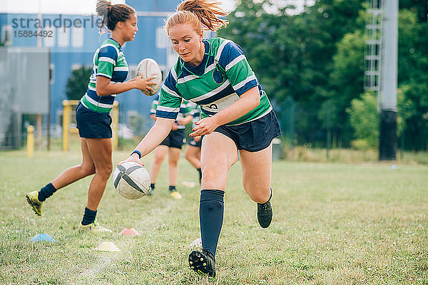 Drei Frauen tragen blaue  weiße und grüne Rugbyhemden auf einem Trainingsplatz  eine läuft mit einem Rugbyball.