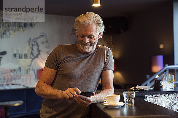 Ein Mann lehnt an einer Bar neben einer Kaffeetasse  lächelt und schaut auf ein Mobiltelefon.