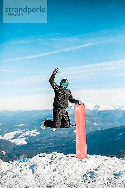 Eine Person  die einen schwarzen Skianzug  einen Helm und eine Schutzbrille trägt und vor Freude auf einem roten Snowboard auf einen Berg springt.