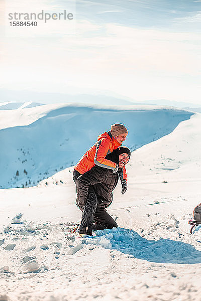 Ein Mann in einem Skianzug  der im Schnee kniet und einer Frau einen Huckepack gibt  lacht und schaut in die Kamera.